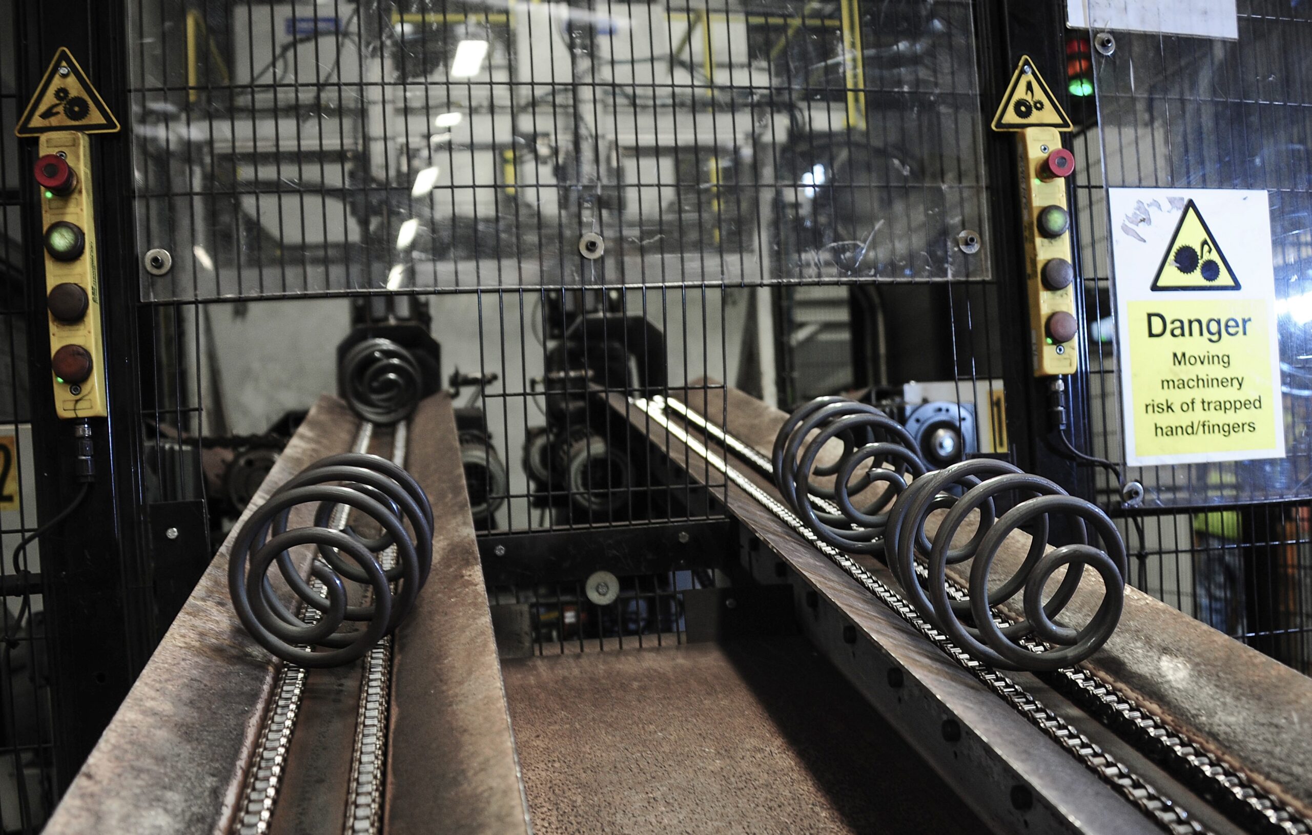 Factory conveyor system transporting large metal coil springs, with safety warning signs and emergency stop buttons visible on a protective cage around moving machinery.